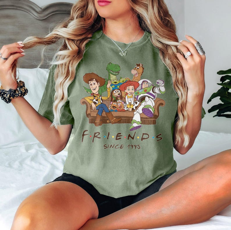 May include: Sage green t-shirt featuring a graphic of Toy Story characters on a brown couch. The word "FRIENDS" is printed in the style of the TV show, with "SINCE 1995" below.