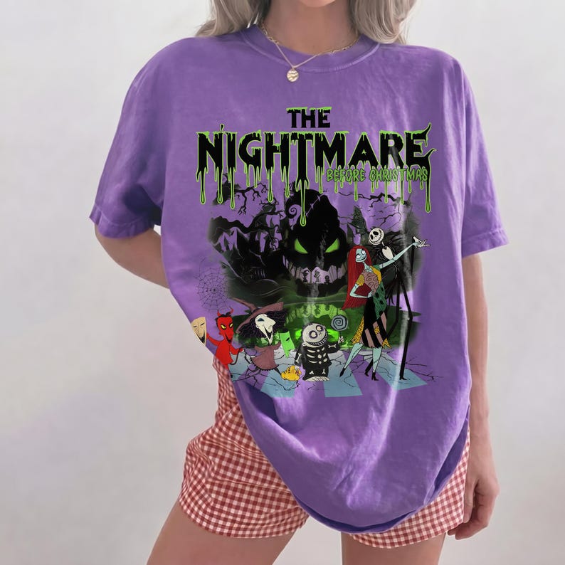 May include: A purple t-shirt featuring a graphic from "The Nightmare Before Christmas." The design includes the text "The Nightmare Before Christmas" in green dripping font, along with characters like Jack Skellington and Sally.