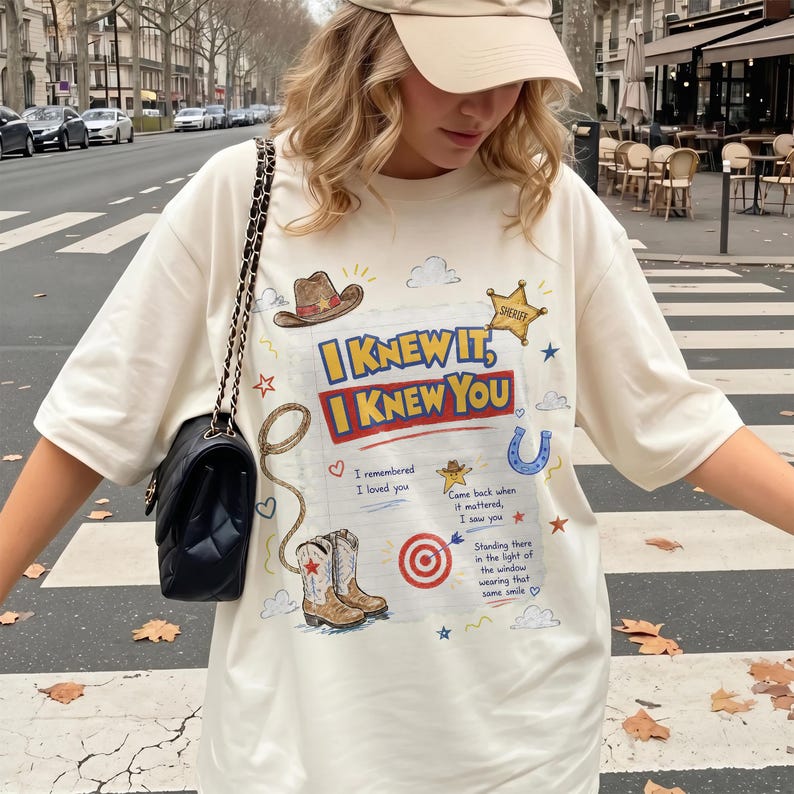 May include: An off-white short-sleeve t-shirt featuring a graphic print with the text "I KNEW IT, I KNEW YOU" alongside illustrations of a cowboy hat, a sheriff star, a lasso, a horseshoe, a target, and cowboy boots on a lined paper background.