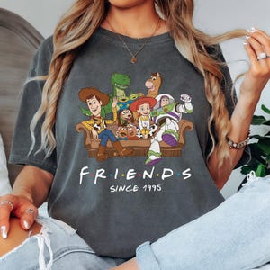 May include: Dark gray t-shirt featuring a graphic of Toy Story characters sitting on a couch. The word "FRIENDS" is printed in white, with "SINCE 1995" below it. The shirt has a crew neck and short sleeves.