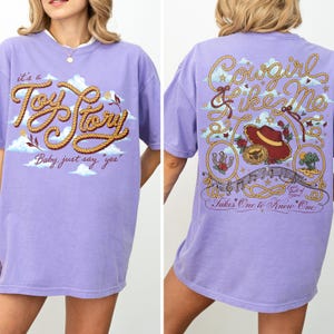 Op de afbeelding: Een lichtpaars t-shirt met een afbeelding op de voorkant met touwachtige tekst "it's a Toy Story Baby, just say "yes"" en een afbeelding op de achterkant met touwranden, een cowboyhoed, een riemgesp en de tekst "Cowgirl like Me".