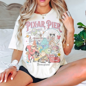 May include: Cream-coloured t-shirt featuring a colourful graphic of Pixar characters and the words "Pixar Pier" and "Disneyland" in a vintage style. The design includes characters from various Pixar films.