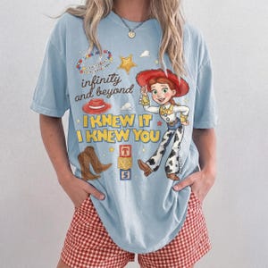 Pode incluir: Uma t-shirt azul-clara com uma estampa gr&aacute;fica de uma cowgirl, um chap&eacute;u vermelho, botas de cowboy castanhas, uma estrela amarela e blocos de alfabeto. O texto diz "infinity and beyond", "I KNEW IT I KNEW YOU" e "YOU'VE GOT A FRIEND IN ME".
