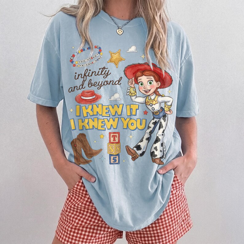 Pode incluir: Uma t-shirt azul-clara com uma estampa gr&aacute;fica de uma cowgirl, um chap&eacute;u vermelho, botas de cowboy castanhas, uma estrela amarela e blocos de alfabeto. O texto diz "infinity and beyond", "I KNEW IT I KNEW YOU" e "YOU'VE GOT A FRIEND IN ME".