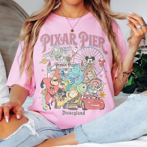 May include: A pink t-shirt with a vibrant graphic of Pixar Pier characters and the Disneyland logo. The design includes characters from Toy Story, Monsters, Inc., and Cars. The text "PIXAR PIER" is arched across the top of the design.