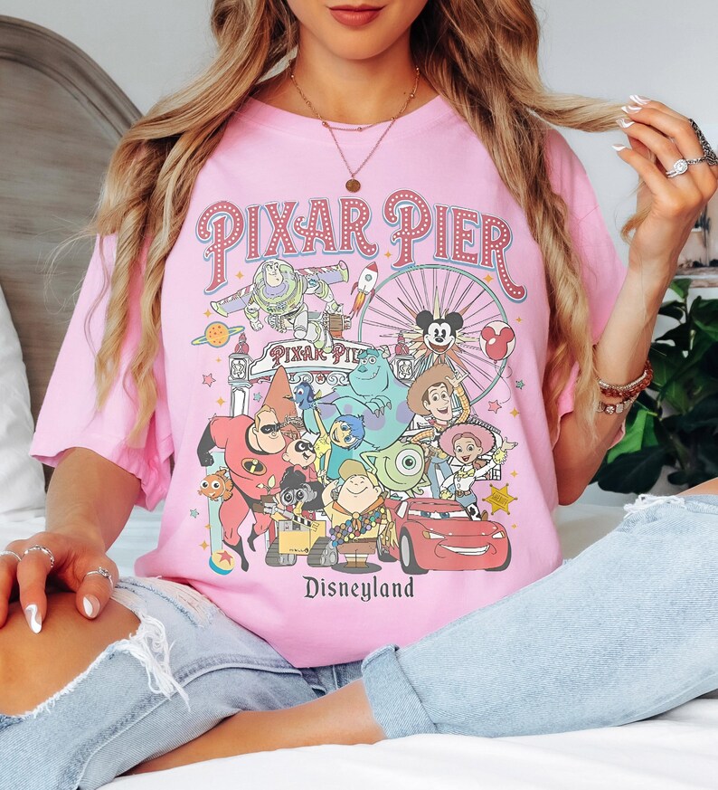 May include: A pink t-shirt with a vibrant graphic of Pixar Pier characters and the Disneyland logo. The design includes characters from Toy Story, Monsters, Inc., and Cars. The text "PIXAR PIER" is arched across the top of the design.