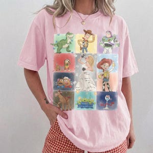K&ouml;nnte beinhalten: Ein hellrosa T-Shirt mit kurzen &Auml;rmeln und einem Raster aus farbenfrohen Illustrationen verschiedener Zeichentrickfiguren, darunter ein gr&uuml;ner Dinosaurier, ein Cowboy, ein Weltraum-Ranger, ein Hund, eine Kartoffelfigur, ein Cowgirl, ein Pferd und Aliens.