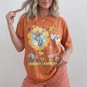 May include: An orange short-sleeve t-shirt featuring a graphic print with a gold ornate frame, a cowboy boot, a star with the text INFINITY AND BEYOND, a beaded bracelet with the text YOU'VE GOT A FRIEND IN ME, and the text I KNEW IT, I KNEW YOU.
