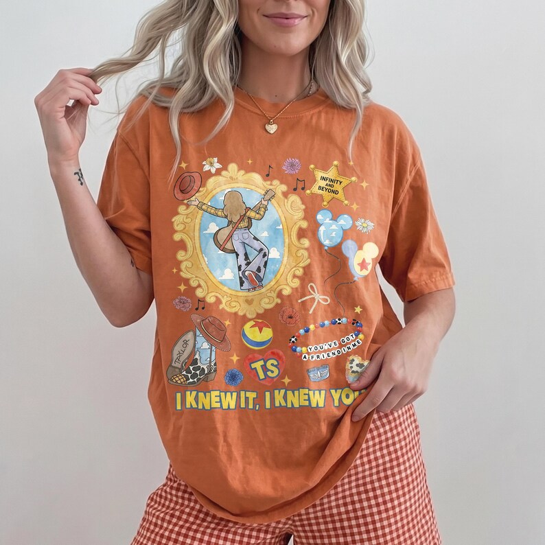 May include: An orange short-sleeve t-shirt featuring a graphic print with a gold ornate frame, a cowboy boot, a star with the text INFINITY AND BEYOND, a beaded bracelet with the text YOU'VE GOT A FRIEND IN ME, and the text I KNEW IT, I KNEW YOU.