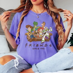 May include: A purple t-shirt featuring a graphic of characters from the movie Toy Story sitting on a couch. The word "FRIENDS" is printed below the graphic, with "SINCE 1995" underneath.