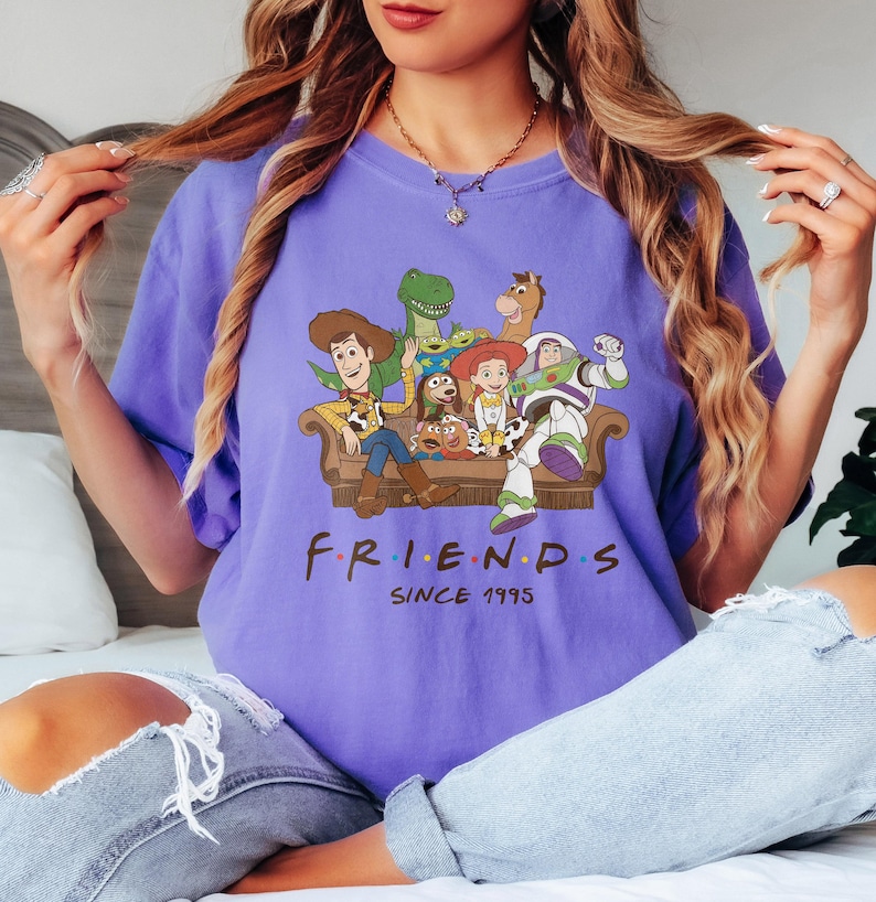 May include: A purple t-shirt featuring a graphic of characters from the movie Toy Story sitting on a couch. The word "FRIENDS" is printed below the graphic, with "SINCE 1995" underneath.