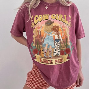 May include: A maroon short-sleeve t-shirt featuring a graphic print of two figures in cowboy hats standing in a desert landscape with cacti and roses. The text reads COW GIRL I KNEW IT I KNEW YOU LIKE ME. The design includes a guitar and western attire.
