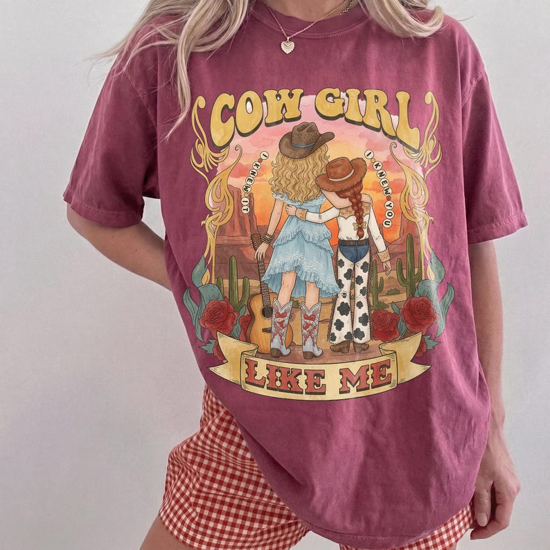 May include: A maroon short-sleeve t-shirt featuring a graphic print of two figures in cowboy hats standing in a desert landscape with cacti and roses. The text reads COW GIRL I KNEW IT I KNEW YOU LIKE ME. The design includes a guitar and western attire.