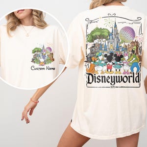 May include: Cream-colored t-shirt featuring a colorful Disney World graphic on the back, including a castle, characters, and the text "Disneyworld 1971." A smaller graphic with the text "Custom Name" is on the front.