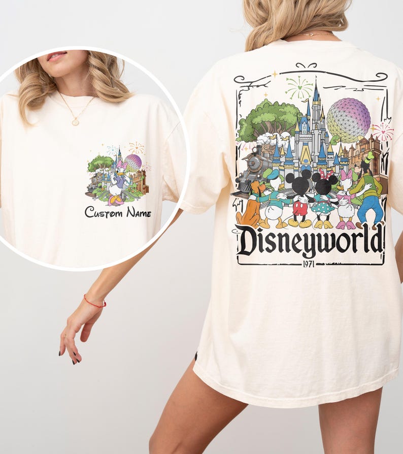 May include: Cream-colored t-shirt featuring a colorful Disney World graphic on the back, including a castle, characters, and the text "Disneyworld 1971." A smaller graphic with the text "Custom Name" is on the front.
