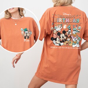 Puede incluir: Una camiseta naranja con personajes de Disney. La parte delantera tiene una caricatura de Goofy con el nombre "MARK". La parte trasera muestra "Disney's Very Happy Birthday" con Mickey, Minnie, Donald, Daisy y Goofy. La camiseta tiene cuello redondo y mangas cortas.