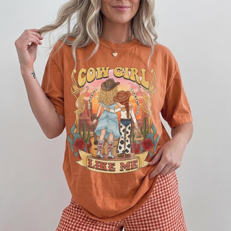 May include: An orange short-sleeved t-shirt featuring a graphic illustration of two figures in cowboy hats standing in a desert landscape with cacti and red roses. The text reads COW GIRL LIKE ME with I KNEW YOU I KNEW YOU written in small letters.