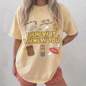 May include: A pale yellow short-sleeve t-shirt featuring the text "I KNEW IT I KNEW YOU" in yellow with a brown outline. Graphics include a beaded necklace, "infinity and beyond" text, a star, clouds, cowboy boots, alphabet blocks, and a red hat.