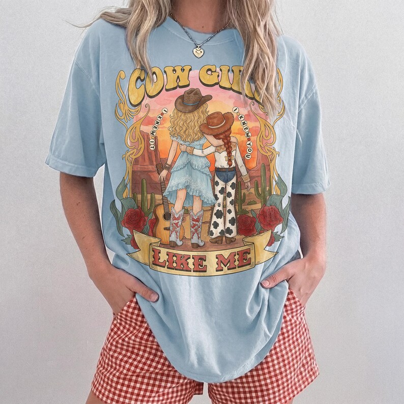 May include: A light blue t-shirt featuring a graphic illustration of two figures in cowboy hats standing in a desert landscape with cacti and red roses. The text reads COW GIRL I KNEW IT I KNEW YOU LIKE ME. The figures wear western attire.