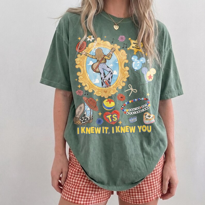 May include: A sage green short-sleeve t-shirt featuring a central gold ornate frame containing a figure with a guitar, surrounded by various icons including a cowboy hat, a ball, a beaded bracelet, and the text "I KNEW IT, I KNEW YOU".