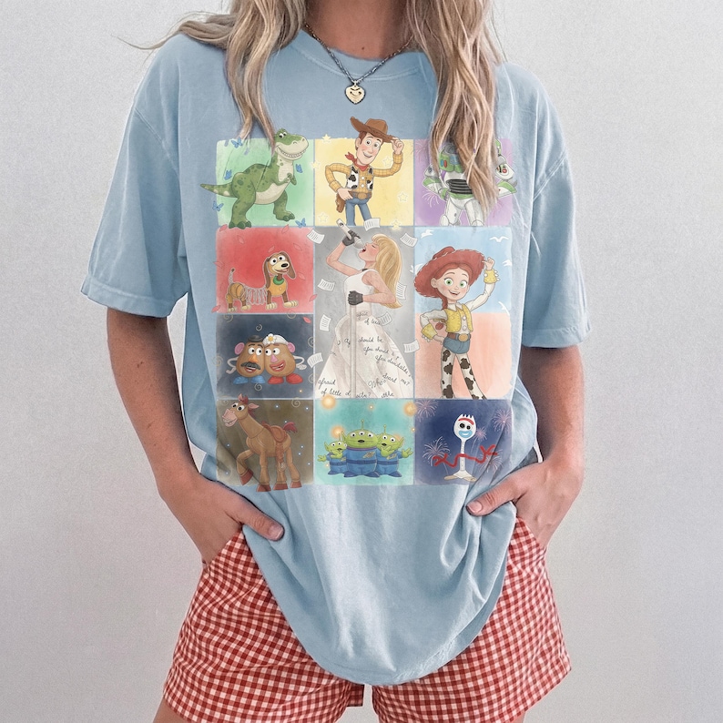 K&ouml;nnte beinhalten: Ein hellblaues Kurzarm-T-Shirt mit einem Raster aus farbenfrohen Illustrationen verschiedener Zeichentrickfiguren, darunter ein Cowboy, ein Weltraum-Ranger, ein Dinosaurier, ein Cowgirl, eine Gabel und andere, mit einer blonden S&auml;ngerin in der Mitte.