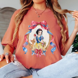 May include: A coral-colored t-shirt featuring a framed illustration of Snow White holding an apple and a basket of flowers, with a deer beside her. The frame is adorned with pink and blue ribbons and bows. The shirt has short sleeves.