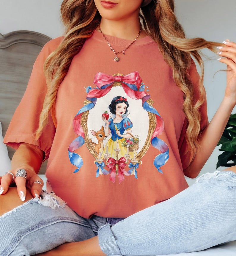 May include: A coral-colored t-shirt featuring a framed illustration of Snow White holding an apple and a basket of flowers, with a deer beside her. The frame is adorned with pink and blue ribbons and bows. The shirt has short sleeves.