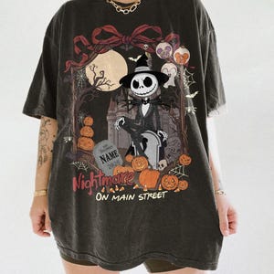 May include: A charcoal gray oversized t-shirt featuring a graphic print of a skeleton character in a suit and hat sitting on a tombstone. The design includes a full moon, a castle, pumpkins, spiderwebs, and the text Nightmare ON MAIN STREET.