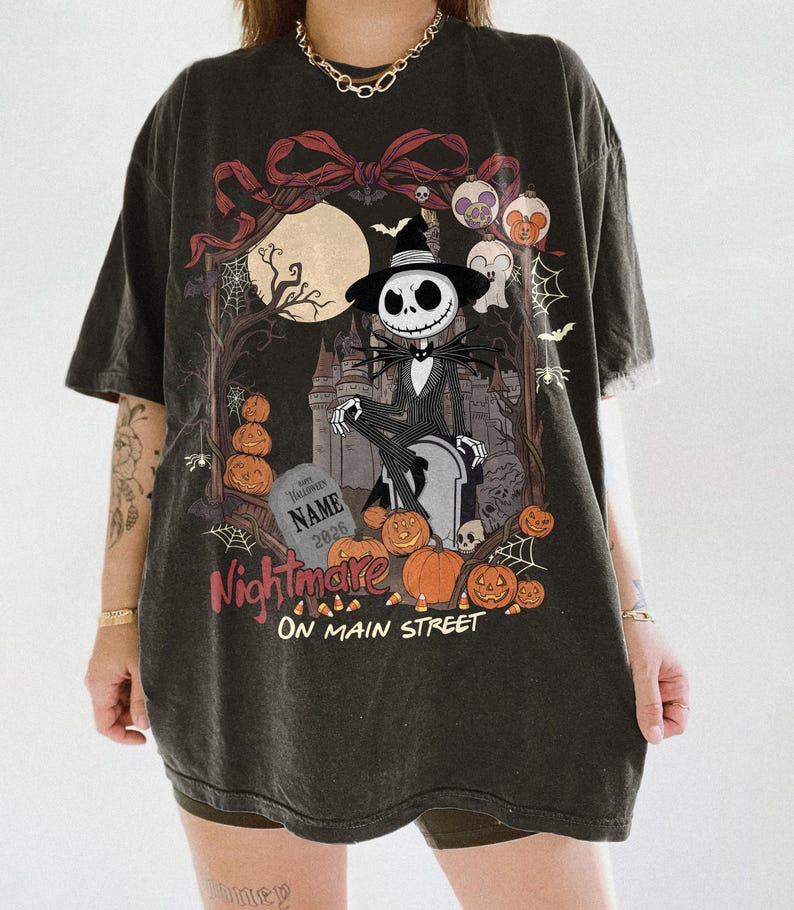 May include: A charcoal gray oversized t-shirt featuring a graphic print of a skeleton character in a suit and hat sitting on a tombstone. The design includes a full moon, a castle, pumpkins, spiderwebs, and the text Nightmare ON MAIN STREET.
