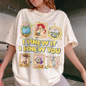 May include: A cream-colored t-shirt featuring a graphic print of Toy Story characters in postage stamp-style frames. The text reads I KNEW IT I KNEW YOU in bold yellow letters with blue outlines. Characters include Jessie, Buzz Lightyear, Woody, and others.