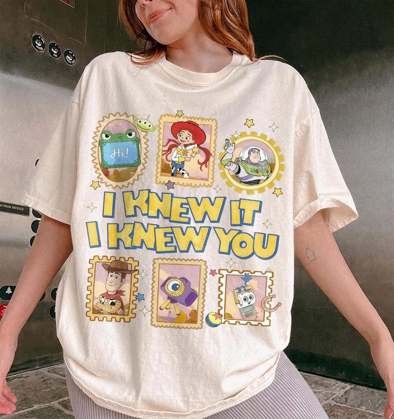 May include: A cream-colored t-shirt featuring a graphic print of Toy Story characters in postage stamp-style frames. The text reads I KNEW IT I KNEW YOU in bold yellow letters with blue outlines. Characters include Jessie, Buzz Lightyear, Woody, and others.