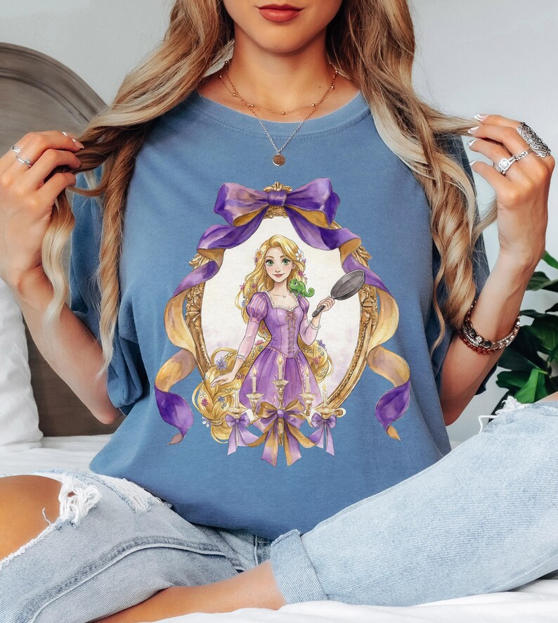 May include: A light blue t-shirt featuring a framed illustration of a princess in a purple dress holding a frying pan. The frame is gold with purple and gold ribbons. The princess has long blonde hair and a green chameleon on her shoulder.