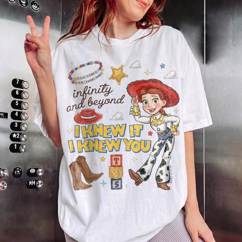 Pode incluir: Uma t-shirt branca com uma impress&atilde;o gr&aacute;fica de uma personagem cowgirl com cabelo ruivo, um chap&eacute;u vermelho e cal&ccedil;as com padr&atilde;o de manchas de vaca. O design inclui o texto "infinity and beyond", "I KNEW IT", "I KNEW YOU", um chap&eacute;u vermelho, botas de cowboy castanhas e blocos de alfabeto.