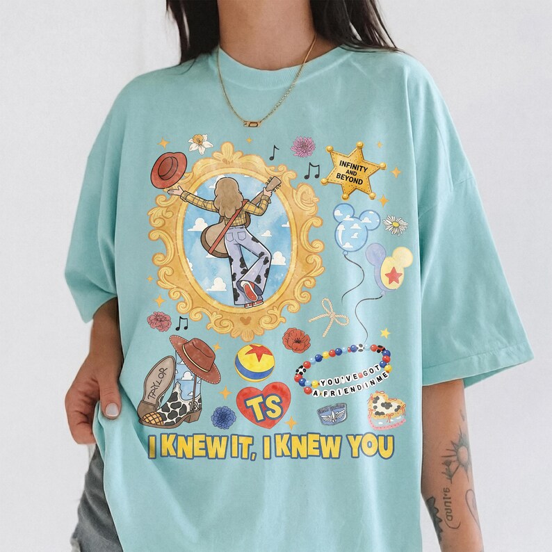 May include: A light blue t-shirt featuring a graphic print of a gold ornate frame containing a person with a guitar, a star with the text INFINITY AND BEYOND, a beaded bracelet with the text YOU'VE GOT A FRIEND IN ME, and the text I KNEW IT, I KNEW YOU.