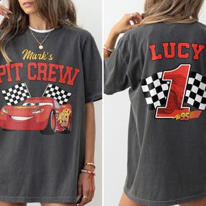 Op de afbeelding: Donkergrijs T-shirt met een rode cartoon raceauto en de tekst "Mark's PIT CREW" op de voorkant. De achterkant van het shirt heeft de naam "LUCY" en het nummer "1" met geblokte vlaggen. Het nummer "95" is ook zichtbaar.