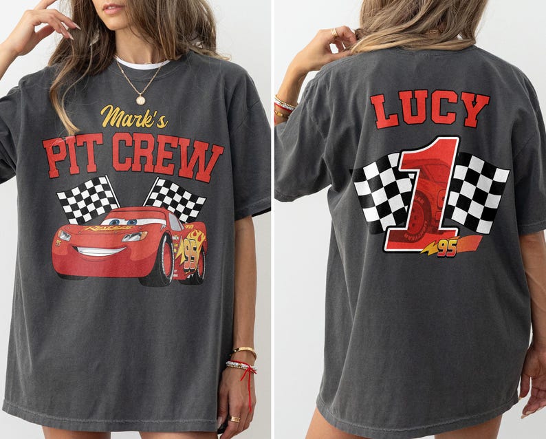 Op de afbeelding: Donkergrijs T-shirt met een rode cartoon raceauto en de tekst "Mark's PIT CREW" op de voorkant. De achterkant van het shirt heeft de naam "LUCY" en het nummer "1" met geblokte vlaggen. Het nummer "95" is ook zichtbaar.