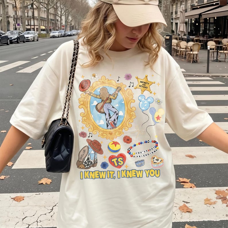 May include: An off-white t-shirt featuring a graphic print of a person playing a guitar inside a gold frame, surrounded by illustrations of a cowboy boot, a heart with the letters TS, balloons, and the text "INFINITY AND BEYOND" and "I KNEW IT, I KNEW YOU."
