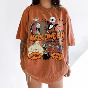 K&ouml;nnte beinhalten: Ein orangefarbenes T-Shirt mit kurzen &Auml;rmeln und einem Grafikdruck von Charakteren aus Nightmare Before Christmas. Das Design enth&auml;lt den Text Nightmare Before HALLOWEEN Trick or Treat Candy Co. SUGARY SURPRISES und SWEET SCARES.