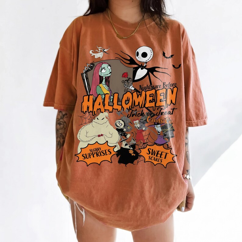 K&ouml;nnte beinhalten: Ein orangefarbenes T-Shirt mit kurzen &Auml;rmeln und einem Grafikdruck von Charakteren aus Nightmare Before Christmas. Das Design enth&auml;lt den Text Nightmare Before HALLOWEEN Trick or Treat Candy Co. SUGARY SURPRISES und SWEET SCARES.
