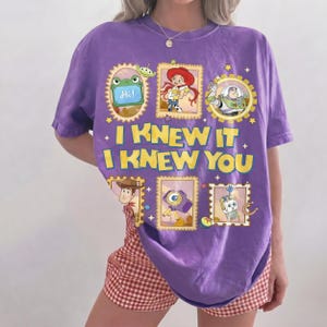 May include: A purple short-sleeve t-shirt featuring a graphic print of various Toy Story characters in framed boxes. The text "I KNEW IT I KNEW YOU" is printed in large yellow letters in the center. The design includes images of Rex, Jessie, Buzz Lightyear, Woody, a camera, and a small robot.