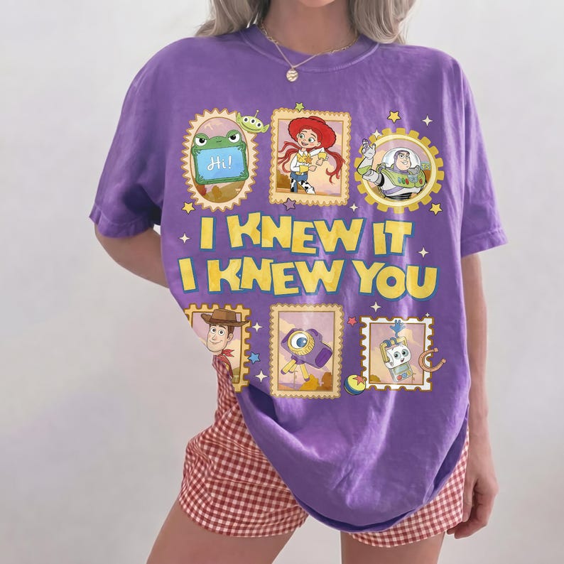 May include: A purple short-sleeve t-shirt featuring a graphic print of various Toy Story characters in framed boxes. The text "I KNEW IT I KNEW YOU" is printed in large yellow letters in the center. The design includes images of Rex, Jessie, Buzz Lightyear, Woody, a camera, and a small robot.