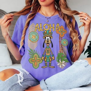 May include: A lilac t-shirt with the word "ALOHA" in yellow lettering. The shirt features a cartoon dog in tribal clothing. The design includes geometric patterns, tropical leaves, and flowers. The shirt is worn by a person.