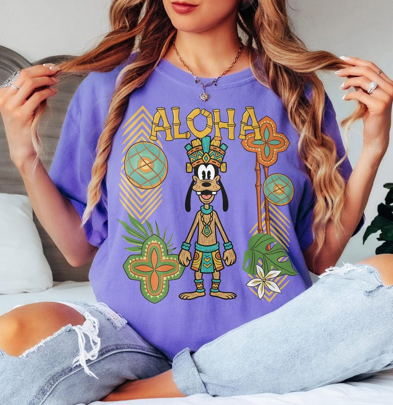 May include: A lilac t-shirt with the word "ALOHA" in yellow lettering. The shirt features a cartoon dog in tribal clothing. The design includes geometric patterns, tropical leaves, and flowers. The shirt is worn by a person.