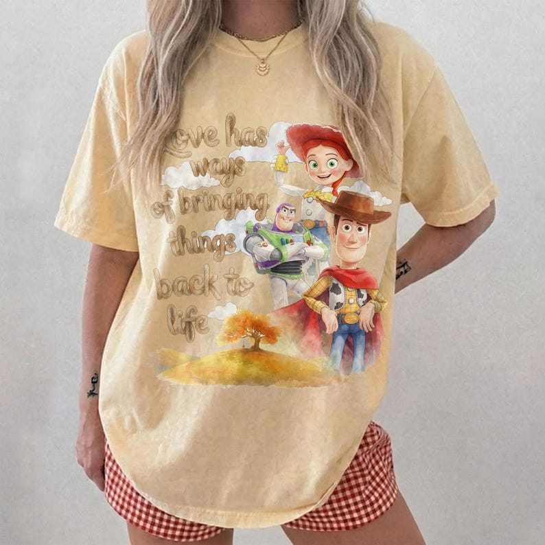 Pu&ograve; includere: Una t-shirt a maniche corte giallo pallido con una stampa grafica di Woody, Buzz Lightyear e Jessie di Toy Story. Il testo recita, Love has ways of bringing things back to life, accompagnato da un'illustrazione di un albero su una collina.