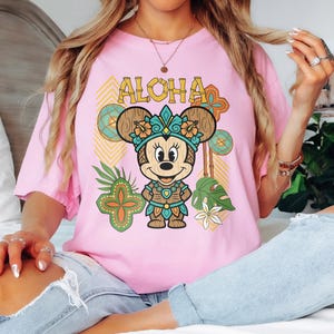 May include: A pale pink t-shirt with a cartoon Minnie Mouse in a Hawaiian outfit. The word "ALOHA" is printed above the character. The design includes tropical flowers, leaves, and geometric patterns in a vibrant style.