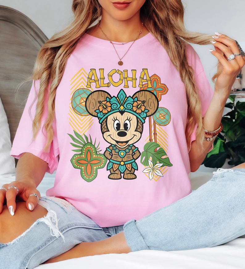 May include: A pale pink t-shirt with a cartoon Minnie Mouse in a Hawaiian outfit. The word "ALOHA" is printed above the character. The design includes tropical flowers, leaves, and geometric patterns in a vibrant style.