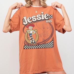 May include: Orange t-shirt featuring the character Jessie from Toy Story. The design includes the name "Jessie" in a stylized font, the year "1995", and a cartoon image of Jessie with a horseshoe and checkered pattern.
