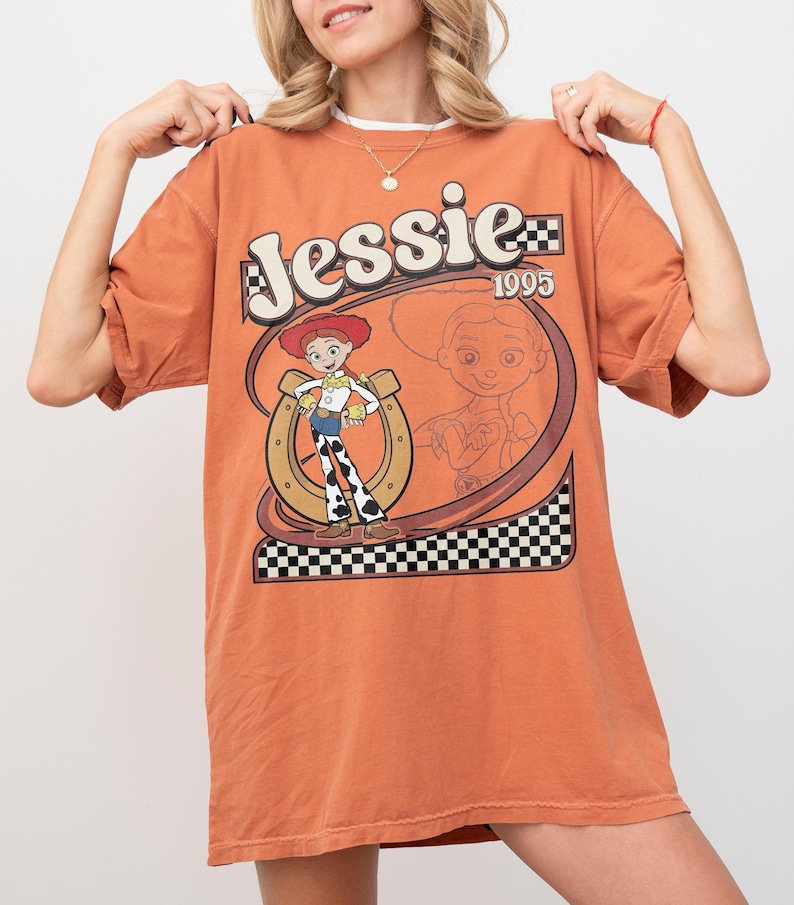 May include: Orange t-shirt featuring the character Jessie from Toy Story. The design includes the name "Jessie" in a stylized font, the year "1995", and a cartoon image of Jessie with a horseshoe and checkered pattern.