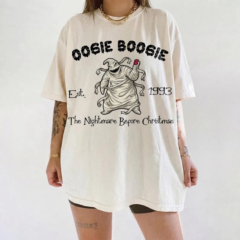 Op de afbeelding: Cr&egrave;mekleurig T-shirt met een afbeelding van Oogie Boogie uit "The Nightmare Before Christmas". Het ontwerp bevat de tekst "Oogie Boogie", "Est. 1993" en "The Nightmare Before Christmas". Het shirt heeft korte mouwen en een relaxte pasvorm.