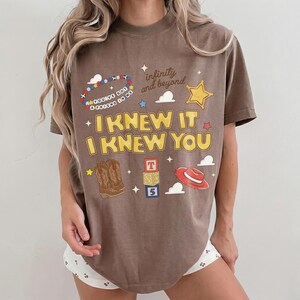 May include: A brown short-sleeve t-shirt featuring a graphic print with the text "I KNEW IT I KNEW YOU", "infinity and beyond", and "YOU'VE GOT A FRIEND IN ME". The design includes illustrations of a beaded bracelet, a star, cowboy boots, blocks, and a hat.